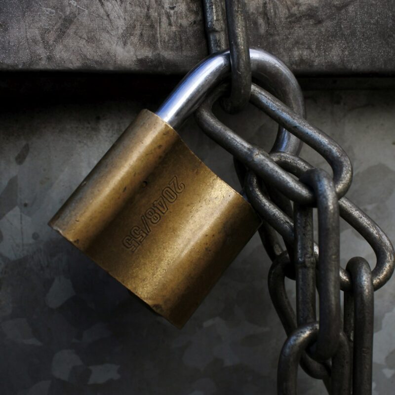 Brass padlock hanging on a steel chain against dark galvanized metal