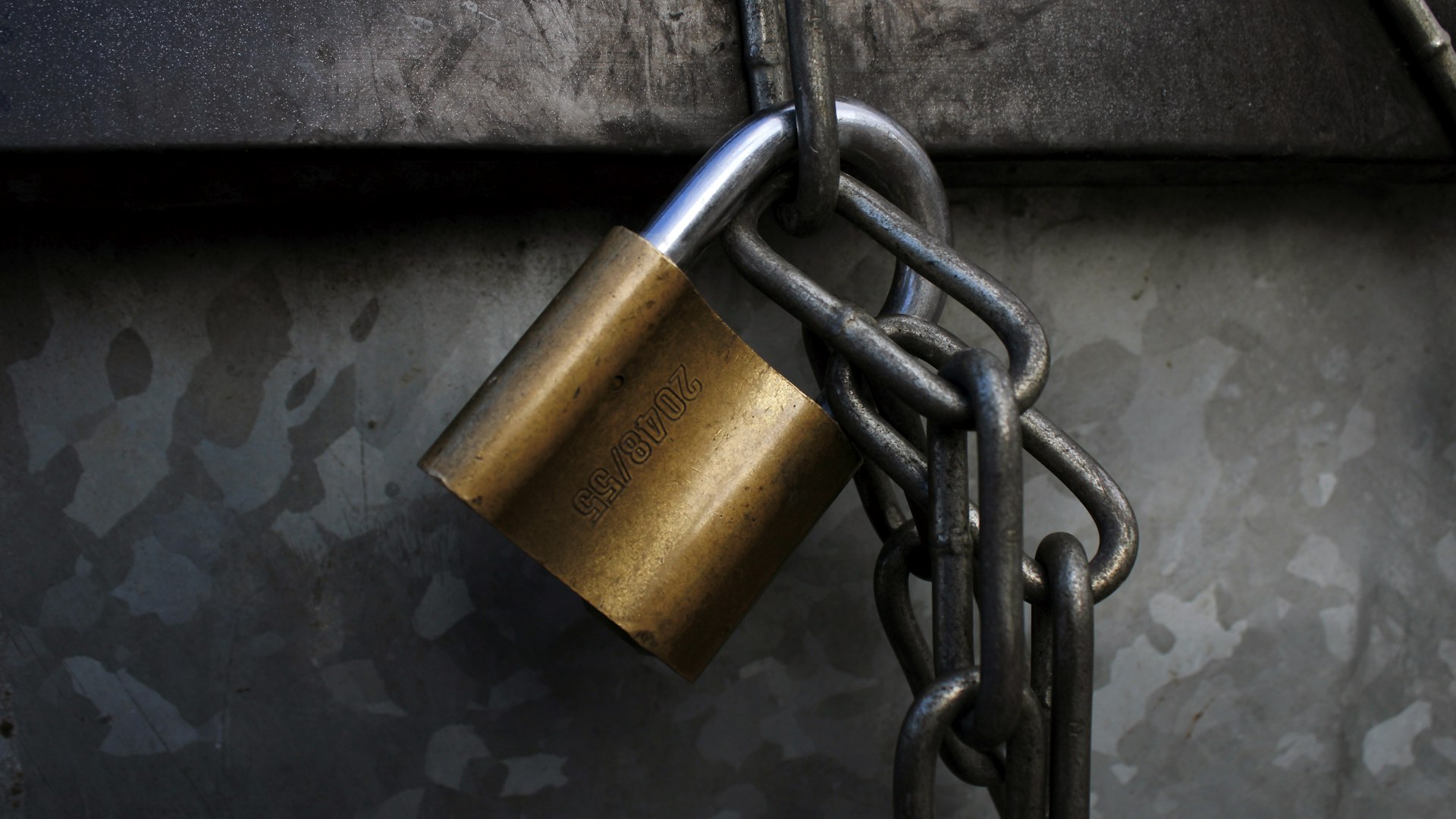 Brass padlock hanging on a steel chain against dark galvanized metal